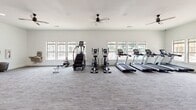 This single-floor fitness center offers a spacious, thoughtfully designed environment for a comprehensive workout experience. The facility is characterized by crisp white walls, neutral-toned carpet flooring, and an abundance of natural light streaming in through multiple large windows that frame outdoor views on multiple sides of the room.

The cardio area is generously outfitted with a lineup of treadmills, elliptical trainers, a stair climber, and a rowing machine, all arranged to take advantage of the natural light and outdoor views. Ceiling fans are mounted overhead, ensuring comfortable airflow throughout the space during workouts.

A dedicated strength training section features a power rack and a multi-functional cable machine, complemented by a full-length wall mirror that visually expands the space and supports proper form during exercise. The open layout allows ample room between equipment, providing a comfortable and unobstructed workout environment.

The facility is accessible via a set of double glass doors at the main entrance, as well as a secondary door providing additional access to an outdoor area. Safety features including exit signage and a fire extinguisher are clearly positioned throughout. The overall ambiance is clean, modern, and well-maintained, making this fitness center a functional and inviting space for users of all fitness levels.