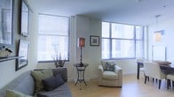This single-floor space offers a bright and airy living area defined by its modern, minimalist design. Large windows fitted with horizontal blinds line the walls, allowing generous amounts of natural light to filter in while offering views of the surrounding urban environment. The light hardwood flooring adds warmth to the otherwise crisp, neutral color palette of white walls and contemporary furnishings. The living area is thoughtfully arranged, featuring a plush sofa with accent pillows in soft greens and grays, complemented by a cream-toned armchair adorned with a patterned throw pillow. A sleek wrought-iron accent table with an elegant table lamp serves as a central decorative element, adding a touch of sophistication to the space. A framed artwork piece on the wall contributes a subtle visual focal point. The corner placement of the windows creates an open, expansive feel, enhancing the sense of space throughout the room. The overall ambiance is clean, calm, and well-lit, making it a comfortable and inviting environment suited for both relaxing and entertaining.