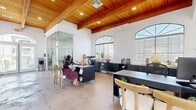 Located at S Monterey Point Condos in Mesa, Arizona, this single-floor office space offers a thoughtfully designed environment that blends professional functionality with refined architectural character. The space is defined by its striking exposed wood-beam ceilings, which lend warmth and a sense of height to the open interior. Polished concrete floors run throughout, providing a clean, modern foundation that complements the crisp white walls.

A series of arched, fan-topped windows line the exterior walls, flooding the space with abundant natural light and offering views of the surrounding outdoor environment. The main work area features sleek dark-toned desks and cabinetry, creating a professional reception and workstation setup suitable for staff and visitors alike. A full-height glass partition elegantly separates a private office or conference area, maintaining visual openness while providing acoustic privacy.

Toward the front of the space, a welcoming lounge area is furnished with plush neutral-toned sofas and armchairs arranged around a central coffee table, anchored by an elegant chandelier overhead. Additional computer workstations are positioned along the walls, offering practical workspace for multiple users. A kitchenette and double-door entry further enhance the utility of this well-appointed office.