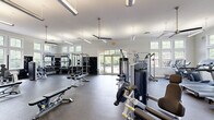 Located at 101 Legacy Terrace in Chapel Hill, NC, this fitness center offers a generously sized, single-floor layout designed to accommodate a full range of workout activities. The space features soaring ceilings that contribute to an open, airy atmosphere, enhanced by large multi-pane windows lining the walls and French doors that flood the interior with natural light and provide views of the surrounding outdoor greenery.

The flooring throughout is covered in durable rubber gym matting, suitable for both cardio and strength training activities. Ceiling fans and pendant lighting fixtures are suspended from the high ceilings, ensuring proper air circulation and visibility across the entire floor.

The fitness center is comprehensively equipped with a dedicated cardio zone featuring treadmills, recumbent bikes, and upright cycling machines. The strength training area includes cable and pulley machines, multi-use weight stations, and selectorized equipment for targeted muscle training. A free weights section offers a full rack of dumbbells, adjustable benches, and a power rack, complemented by large wall mirrors that allow users to monitor their form. A flat-screen television is mounted on the wall for added convenience. French doors on one side of the space open to an outdoor area, providing easy access and a seamless connection to the exterior.