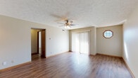 This single-floor residence offers a thoughtfully designed layout that balances comfort and functionality throughout. The living area is a standout space, featuring rich hardwood-style flooring, a ceiling fan with integrated lighting, and a large sliding glass door framed by vertical blinds that floods the room with natural light. A charming octagonal accent window adds a distinctive architectural touch to the living space. The open-concept design connects the living area seamlessly to the kitchen, which is equipped with warm oak cabinetry, dark granite-style countertops, black appliances including a refrigerator and dishwasher, and hardwood-style flooring that continues the cohesive aesthetic. The bedrooms are carpeted for comfort, with neutral walls and standard window blinds providing natural light and privacy. One bedroom notably features two sliding wood-framed closets, offering generous storage capacity. The bathroom is well-appointed with a full tub and shower combination, a dark granite-topped vanity with rich wood cabinetry, tile-style flooring, and clean, classic fixtures. Textured ceilings are present throughout the home, adding subtle character, while consistent warm-toned trim and baseboards unify the overall aesthetic of the space.