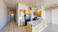 Located in the heart of Dayton, Ohio, this loft-style unit at 500 E Third St blends industrial character with modern functionality. The open-concept living and kitchen area is defined by whitewashed exposed brick walls, high ceilings with visible ductwork, and light hardwood-style flooring that flows throughout. The kitchen features warm honey-toned cabinetry, granite-style countertops, black appliances including a refrigerator and range, a deep sink, and a peninsula island that doubles as a breakfast bar — all illuminated by Edison-bulb pendant lights suspended from industrial track railings overhead. Large windows in the living area flood the space with natural light and offer views of the surrounding Dayton cityscape. The hallway, lined with striking exposed brick, leads to the spacious bedroom, which is carpeted for comfort and features exposed ductwork overhead and a sliding barn-style door. A dedicated closet with built-in shelving and hanging space provides ample storage. Two full bathrooms serve the unit, one featuring a full tub/shower combo with granite-style vanity countertops and wood cabinetry. The overall ambiance is distinctly urban and loft-inspired, with a thoughtful balance of raw architectural elements and comfortable living finishes.