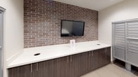 This mail room is designed with both functionality and aesthetic appeal in mind. The space is anchored by multiple banks of floor-mounted, aluminum individual mailboxes lining one wall, providing ample mail storage capacity for a large number of residents or occupants. Recessed ceiling lighting illuminates the room evenly, creating a clean and welcoming environment.

A standout design feature is the textured brick accent wall in warm brown tones, which adds visual depth and character to the space. Set against this wall is a wall-mounted flat-screen display, positioned above a built-in countertop with dark wood-grain cabinetry below. The counter spans the length of the accent wall and includes integrated cable management cutouts, making it a practical surface for sorting mail and packages.

A central island-style table with a white surface provides additional workspace in the middle of the room, offering a convenient area for handling deliveries. The flooring consists of large-format neutral tiles, contributing to the room's clean, modern appearance. A solid entry door with a small window provides secure access to the space. Overall, the mail room balances practicality with polished design details, creating an efficient and pleasant environment for daily use.