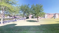 This single-floor property is set within a generously sized outdoor environment that emphasizes open space, greenery, and a welcoming atmosphere. The grounds are defined by a well-kept, expansive lawn that stretches across the property, offering a clean and manicured appearance throughout. Mature deciduous trees are thoughtfully distributed across the landscape, providing natural shade and contributing to the park-like character of the space. Their full canopies create a pleasant, tree-lined setting that enhances both the aesthetic and comfort of the outdoor areas. The building itself features classic brick exterior construction, lending a sense of solidity and timeless architectural character. Paved walkways and a parking area are visible along the periphery, providing practical accessibility while maintaining the openness of the green space. The overall ambiance is calm and well-organized, with the combination of natural landscaping and structured hardscaping creating a balanced environment. Under clear, sunny skies, the property benefits from abundant natural light that highlights the vibrancy of the green lawn and the warm tones of the brick facades, making it an inviting and functional space.