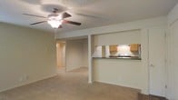 This single-floor residence offers an open-concept layout that connects the living area to the kitchen, creating a cohesive and functional living environment. The living space is finished with wall-to-wall carpeting in a warm beige tone, contributing to a comfortable and understated aesthetic. A ceiling fan with an integrated light fixture is centrally positioned overhead, providing both air circulation and ambient lighting. The textured popcorn ceiling adds a classic residential character to the space.

The kitchen is accessible via a raised breakfast bar with a dark countertop, which serves as both a workspace and a casual dining or serving area. Honey-toned wooden cabinetry lines the upper walls, complemented by a white electric range and an under-cabinet range hood. The kitchen design is efficient and straightforward, maximizing utility within a compact footprint.

A structural column near the center of the space subtly defines the transition between the living and kitchen areas without interrupting the open feel. A hallway visible in the background leads to additional rooms, suggesting a practical and well-organized floor plan. Neutral wall colors throughout maintain a clean, move-in-ready appearance.
