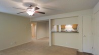 This single-floor residence offers an open-concept layout that connects the living area to the kitchen, creating a cohesive and functional living environment. The living space is finished with wall-to-wall carpeting in a warm beige tone, contributing to a comfortable and understated aesthetic. A ceiling fan with an integrated light fixture is centrally positioned overhead, providing both air circulation and ambient lighting. The textured popcorn ceiling adds a classic residential character to the space.

The kitchen is accessible via a raised breakfast bar with a dark countertop, which serves as both a workspace and a casual dining or serving area. Honey-toned wooden cabinetry lines the upper walls, complemented by a white electric range and an under-cabinet range hood. The kitchen design is efficient and straightforward, maximizing utility within a compact footprint.

A structural column near the center of the space subtly defines the transition between the living and kitchen areas without interrupting the open feel. A hallway visible in the background leads to additional rooms, suggesting a practical and well-organized floor plan. Neutral wall colors throughout maintain a clean, move-in-ready appearance.