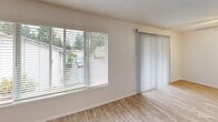 Located in University Place, WA, this single-story unit offers a thoughtfully laid-out floor plan with a living room and one bedroom. The living room is a welcoming, open space finished with light wood-style vinyl flooring and neutral walls that create a clean, airy atmosphere. A large picture window paired with a sliding glass door floods the room with natural light and provides views of the surrounding greenery, making the space feel bright and connected to the outdoors. Vertical blinds and horizontal window blinds offer flexible light control throughout.

The kitchen, visible in the dollhouse view, features gray cabinetry, a range, and efficient use of space, providing a functional cooking area. A hallway with a paneled gray door connects the main living area to the private bedroom.

The bedroom is a comfortably sized retreat, carpeted in neutral tones for warmth underfoot. It includes a built-in closet with a clothing rod for convenient storage, a window that allows natural light in, and a ceiling light fixture. The textured ceilings are consistent throughout the unit, adding a classic residential character. Overall, the space presents a clean, move-in ready environment with a practical and cohesive design.