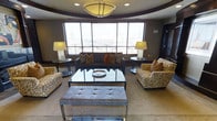 This single-floor space showcases a thoughtfully designed living area that blends contemporary elegance with comfortable functionality. The lounge area is anchored by a large, dark upholstered sofa adorned with decorative pillows in warm amber and gold tones, complemented by two patterned accent chairs featuring bold, abstract detailing. A sleek glass-top coffee table with a metallic frame serves as the focal point of the seating arrangement, while a tufted ottoman provides additional versatility at the room's edge.

A wide, multi-pane window dominates one wall, flooding the space with natural light and offering expansive views of the surrounding urban skyline. The window is fitted with horizontal blinds for adjustable light control. Flanking the sofa, two matching table lamps on round side tables provide warm ambient lighting, supplemented by recessed ceiling fixtures and a centrally positioned flush-mount ceiling light with a frosted diffuser.

The ceiling features a subtle tray detail that adds architectural depth to the room. Neutral-toned carpeting grounds the space, while dark wall accents provide contrast. The overall ambiance is polished, urban, and inviting.