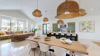 Located at 26502 Longboard Dr in Bonita Springs, Florida, this single-floor community space offers a generous, open-concept layout filled with natural light and modern coastal design. Soaring vaulted ceilings amplify the sense of openness throughout, while large floor-to-ceiling windows and slim vertical accents frame lush tropical views of palm trees and an outdoor pool area.

The interior is anchored by polished light tile flooring that flows seamlessly across multiple functional zones. A comfortable lounge area features a built-in sectional sofa with olive-green base, accented by colorful throw pillows and complemented by woven rattan seating and lush indoor plants, creating a relaxed, resort-inspired atmosphere. A secondary lounge zone offers additional soft seating and curated artwork on crisp white walls.

The dining area centers around a large natural wood communal table surrounded by woven white chairs, illuminated by a striking cluster of oversized woven pendant lights that add warmth and texture overhead. Adjacent to the dining space, a bar counter with deep green cabinetry and bar-height seating provides a functional and stylish gathering point.

A sliding barn door reveals an additional private room, while the overall palette of white, sage green, and warm neutrals delivers a cohesive, inviting ambiance throughout the space.