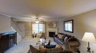 This two-story home offers a comfortable and well-appointed living space with thoughtful design details throughout. The living room serves as the centerpiece of the main floor, featuring a tiled fireplace with a classic white mantel that anchors the room as a focal point. Crown molding runs along the upper walls, adding a refined architectural touch, while a textured ceiling and ceiling fan contribute to the room's layered character.

Natural light filters in through multiple windows dressed with sheer curtains, brightening the neutral, warm-toned walls and light-colored carpeting. The generous layout of the living room easily accommodates a large sectional sofa arrangement, a glass coffee table, and a dedicated reading nook near the front windows. A decorative mirror on one wall adds depth and visual interest to the space.

The overall ambiance is warm and inviting, with a neutral color palette that provides a versatile backdrop for furnishings and decor. The combination of classic architectural details, such as the crown molding and fireplace surround, with practical features like the ceiling fan, reflects a balance of comfort and style carried throughout this two-floor home.