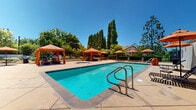 Located in Thousand Oaks, California, this outdoor amenity area offers a well-appointed resort-style retreat designed for relaxation and recreation. The centerpiece is a generously sized swimming pool with crystal-clear turquoise water, accessible via stainless steel entry rails and surrounded by a broad stone-tiled deck that provides ample space for lounging and socializing. Adjacent to the main pool sits a circular spa, perfect for unwinding after a swim.

The pool area is thoughtfully furnished with vibrant orange canopy cabanas, each sheltering cushioned chaise lounges that invite guests to relax in shaded comfort. Additional open-air lounge chairs and umbrella-shaded seating are arranged around the deck, offering both sun and shade options throughout the day. A pergola-covered seating alcove along one side of the space provides a more sheltered gathering area with a cozy, intimate atmosphere.

Tall Italian cypress trees line the perimeter, lending a sense of privacy and a distinctly Mediterranean character to the space. Lush greenery, manicured hedges, and colorful landscaping frame the entire area, creating a serene, garden-like environment. The surrounding buildings feature a warm stucco exterior with tile rooflines that complement the outdoor aesthetic. Under the consistently clear Southern California sky, this outdoor space delivers an inviting and polished recreational experience.