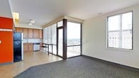 This single-floor space showcases an open-concept layout that seamlessly connects the living area and kitchen. The most striking feature is the expansive floor-to-ceiling window system framed in dark trim, which floods the interior with natural light and offers panoramic elevated views of the surrounding cityscape. The windows are fitted with horizontal blinds for adjustable light control and privacy.

The living area is finished with dark carpeting, providing a grounded contrast to the lighter tones throughout the rest of the space. The kitchen occupies a compact yet functional footprint, featuring warm wood cabinetry, light countertops, and stainless steel appliances including a dishwasher. A narrow high-top bar table with stools sits at the transition between the kitchen and the windowed area, creating a casual dining or workspace zone. The flooring in the kitchen area is a light-toned hard surface, distinguishing it from the carpeted living space.

The overall ambiance is contemporary and urban, characterized by clean lines, a neutral color palette, and an abundance of natural light. The elevated position of the unit enhances the sense of openness and connection to the city below.