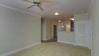 This single-floor space showcases an open-concept design that seamlessly connects the living area to the kitchen. The living room is finished with plush, light-toned carpeting and neutral-painted walls accented by white crown molding and baseboards. A ceiling fan with a light fixture is centrally mounted, providing both comfort and ambient lighting.

The kitchen is positioned to the right and features a raised breakfast bar that serves as a natural divider between the living and cooking areas. Pendant lights hang above the bar, adding a decorative touch while providing task lighting. White cabinetry lines the kitchen, complementing the overall light and airy aesthetic. A refrigerator is visible at the far end of the kitchen, and darker upper cabinetry provides a subtle contrast.

A hallway extends toward the back of the space, leading to additional rooms and finished with durable wood-look flooring that contrasts with the carpeted living area. The transition in flooring clearly delineates functional zones within the layout. The neutral color palette, crown molding details, and consistent finishes throughout contribute to a cohesive and comfortable living environment.