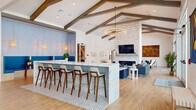 This thoughtfully designed single-floor community clubhouse offers nearly 2,000 square feet of versatile gathering space, blending modern aesthetics with warm, inviting architectural details. Dramatic vaulted ceilings adorned with rustic exposed wood beams define the main social area, creating an airy, open atmosphere bathed in natural light streaming through oversized windows and glass doors.

At the heart of the space is a striking large island clad in marble-look stone and patterned tile, surrounded by mid-century modern bar stools — an ideal spot for casual socializing. Adjacent to the island, a cozy blue-accented nook with built-in bench seating offers a more intimate retreat.

The lounge area features a floor-to-ceiling stacked stone fireplace wall with a mounted flat-screen TV, complemented by stylish blue sofas, accent chairs, and a statement ring chandelier overhead. Curated artwork adds vibrant color throughout.

A long, light-filled entry corridor with shiplap walls, a skylight, a coffee and beverage station, and views to the outdoors creates a welcoming arrival experience. A separate dining room with a large wood table, quilted velvet chairs, a candle-style chandelier, and bold large-scale art opens directly to an outdoor terrace, seamlessly extending the entertaining space.