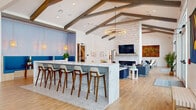 This thoughtfully designed single-floor community clubhouse offers nearly 2,000 square feet of versatile gathering space, blending modern aesthetics with warm, inviting architectural details. Dramatic vaulted ceilings adorned with rustic exposed wood beams define the main social area, creating an airy, open atmosphere bathed in natural light streaming through oversized windows and glass doors.

At the heart of the space is a striking large island clad in marble-look stone and patterned tile, surrounded by mid-century modern bar stools — an ideal spot for casual socializing. Adjacent to the island, a cozy blue-accented nook with built-in bench seating offers a more intimate retreat.

The lounge area features a floor-to-ceiling stacked stone fireplace wall with a mounted flat-screen TV, complemented by stylish blue sofas, accent chairs, and a statement ring chandelier overhead. Curated artwork adds vibrant color throughout.

A long, light-filled entry corridor with shiplap walls, a skylight, a coffee and beverage station, and views to the outdoors creates a welcoming arrival experience. A separate dining room with a large wood table, quilted velvet chairs, a candle-style chandelier, and bold large-scale art opens directly to an outdoor terrace, seamlessly extending the entertaining space.