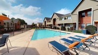 This residential community presents a welcoming outdoor environment centered around a generously sized swimming pool with crystal-clear blue water. The pool area is framed by a tan stone-tiled deck and surrounded by a secure metal fence, offering both safety and an open, airy feel. Multiple blue lounge chairs with orange accents are arranged along the poolside, providing ample space for relaxation and sunbathing. A shaded seating area with an orange umbrella and additional chairs offers a comfortable spot for outdoor gatherings. The community buildings feature a combination of warm brick and neutral-toned horizontal siding exteriors, complemented by pitched rooflines and private balconies. Mature trees and well-tended shrubs, including oleander plantings, add natural greenery throughout the grounds, enhancing the inviting atmosphere. A secure metal lattice gate provides controlled access to the pool and surrounding areas, contributing to the overall safety and privacy of the community. The outdoor spaces are bathed in natural sunlight under open skies, creating a bright and pleasant environment. The thoughtful landscaping and amenity offerings reflect a community designed with resident comfort and outdoor living in mind.