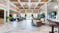 Located at 1025 Retreat Dr in Bartow, Florida, this single-story community clubhouse delivers a refined coastal aesthetic across a generously proportioned floor plan. The grand entrance welcomes visitors with a striking blue geometric area rug and towering shiplap-paneled walls that set an immediately sophisticated tone. The heart of the space is a sweeping open-plan gathering area adorned with large-format marble-look porcelain tile flooring that extends throughout. Soaring coffered ceilings with warm wood inlays, statement chandeliers, and ceiling fans lend architectural drama, while an abundance of natural light pours in through expansive glass doors that frame views of the outdoor pool area. Lush tropical greenery punctuates the interior, reinforcing the Florida resort ambiance. Multiple seating arrangements accommodate both casual lounging and formal meetings, with coordinated furnishings in navy, cream, and warm wood tones. A full kitchen provides functional amenities for events and gatherings. Private office spaces branch off the main hall, each featuring shiplap accent walls, marble-look tile flooring, arched windows, and tastefully appointed wood desks with ample storage. The overall atmosphere is polished yet welcoming, blending upscale design details with practical, community-focused functionality.