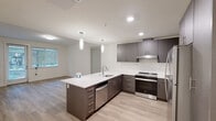 Located at 201–233 S 348th St in Federal Way, Washington, this single-floor unit offers a thoughtfully designed layout with modern finishes throughout. Light-toned luxury vinyl plank flooring runs consistently across the entire space, contributing to a clean, cohesive aesthetic.

The kitchen serves as a central highlight, featuring a large peninsula with a crisp white quartz countertop, an undermount sink, and pendant lighting overhead. Dark charcoal cabinetry with brushed nickel hardware lines both the upper and lower walls, complemented by a subway tile backsplash and stainless steel appliances, including a Whirlpool range, built-in dishwasher, and refrigerator.

The living area flows naturally from the kitchen and receives ample natural light through large windows with horizontal blinds. A glass door provides direct outdoor access, bringing in additional greenery views. The bedrooms are well-proportioned, with multiple windows offering pleasant outdoor sightlines.

The bathrooms are finished in a contemporary style, one featuring a backlit LED vanity mirror, dark cabinetry with integrated storage tower, and light flooring. A second bathroom includes a tub-shower combination with a frosted privacy window. A dedicated laundry area houses a full-size Whirlpool front-loading washer and dryer with overhead shelving.