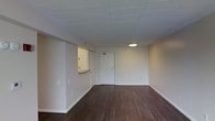 This single-floor space presents a clean, functional layout designed for practical use. The interior is finished with rich, dark hardwood flooring that extends throughout the open main area, providing a warm contrast against the neutral white walls and white drop tile ceiling. The ceiling features a centrally placed flush-mounted circular light fixture, ensuring even ambient illumination across the space.

The main area is notably open and unobstructed, offering flexible use of the floor plan. A partial wall with a pass-through counter extends from one side, creating a subtle division between the main room and an adjacent area, likely a kitchenette or utility space. This architectural detail adds a sense of structure without fully closing off the space. The entry door, situated at the far end of the room, is a standard white panel door with a simple, clean finish. Electrical outlets are distributed along the baseboards throughout the space. The white painted baseboards and trim provide a tidy, cohesive finish throughout, contributing to the overall minimalist and well-maintained character of the interior.