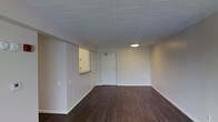 This single-floor space presents a clean, functional layout designed for practical use. The interior is finished with rich, dark hardwood flooring that extends throughout the open main area, providing a warm contrast against the neutral white walls and white drop tile ceiling. The ceiling features a centrally placed flush-mounted circular light fixture, ensuring even ambient illumination across the space.

The main area is notably open and unobstructed, offering flexible use of the floor plan. A partial wall with a pass-through counter extends from one side, creating a subtle division between the main room and an adjacent area, likely a kitchenette or utility space. This architectural detail adds a sense of structure without fully closing off the space. The entry door, situated at the far end of the room, is a standard white panel door with a simple, clean finish. Electrical outlets are distributed along the baseboards throughout the space. The white painted baseboards and trim provide a tidy, cohesive finish throughout, contributing to the overall minimalist and well-maintained character of the interior.