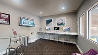 This single-floor community clubhouse is thoughtfully designed to serve as a multifunctional hub, combining a business center, lounge, and leasing office within a cohesive, contemporary layout. The main lounge area showcases rich dark hardwood flooring, recessed lighting, and an open floor plan that creates a welcoming and spacious atmosphere. A comfortable seating area with a sofa and area rug anchors the space, complemented by framed blueprint-style artwork on the walls.

The business center is equipped with multiple iMac workstations arranged along a sleek white countertop with modern bar-height seating. Wall-mounted flat-screen televisions and ocean-themed artwork add visual interest throughout. A glass partition wall elegantly separates the business center from the lounge, maintaining an open feel while defining each zone.

A secondary workstation nook features large windows overlooking a resort-style pool area with palm trees and outdoor seating, bringing in natural light and vibrant outdoor views. The leasing office section includes a glass-top table with bar stools and a dedicated desk area. Patterned carpet in the business center zones contrasts with the hardwood flooring in the lounge, clearly delineating each functional area within this well-appointed space.