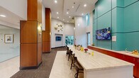 Located at 805 North Olive Avenue in West Palm Beach, Florida, this single-floor amenity space offers a thoughtfully designed and well-appointed environment spanning a generous footprint. The space is characterized by soaring double-height ceilings that create an open, airy atmosphere, complemented by recessed lighting and sleek pendant fixtures that illuminate the interior with a warm, inviting glow.

The main lounge area serves as the centerpiece, featuring a long marble-topped bar counter lined with dark wood barstools — ideal for socializing or working. Bold teal accent wall panels provide a vibrant contrast against the warm wood-clad columns that anchor the space architecturally. Wall-mounted flat-screen televisions are positioned throughout for both informational and entertainment purposes. Decorative gold wall art adds a touch of sophistication.

A seating nook offers small bistro-style tables and chairs alongside floor-to-ceiling sheer curtains, with framed abstract artwork displayed nearby. A glass-walled office or concierge area is visible from the lounge, furnished with teal leather seating and a marble coffee table. A fully equipped kitchen area includes stainless steel appliances, cabinetry, a sink, and a coffee station. The patterned carpet flooring with geometric inlay detailing ties the entire space together cohesively.