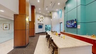 Located at 805 North Olive Avenue in West Palm Beach, Florida, this single-floor amenity space offers a thoughtfully designed and well-appointed environment spanning a generous footprint. The space is characterized by soaring double-height ceilings that create an open, airy atmosphere, complemented by recessed lighting and sleek pendant fixtures that illuminate the interior with a warm, inviting glow.

The main lounge area serves as the centerpiece, featuring a long marble-topped bar counter lined with dark wood barstools — ideal for socializing or working. Bold teal accent wall panels provide a vibrant contrast against the warm wood-clad columns that anchor the space architecturally. Wall-mounted flat-screen televisions are positioned throughout for both informational and entertainment purposes. Decorative gold wall art adds a touch of sophistication.

A seating nook offers small bistro-style tables and chairs alongside floor-to-ceiling sheer curtains, with framed abstract artwork displayed nearby. A glass-walled office or concierge area is visible from the lounge, furnished with teal leather seating and a marble coffee table. A fully equipped kitchen area includes stainless steel appliances, cabinetry, a sink, and a coffee station. The patterned carpet flooring with geometric inlay detailing ties the entire space together cohesively.