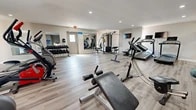Located at 4300 Pan American Fwy in Albuquerque, New Mexico, this single-floor community amenity space is divided into two distinct areas: a fully equipped fitness center and a comfortable lounge and workspace.

The fitness center is generously proportioned, featuring light wood-style flooring, recessed lighting, and large wall mirrors that enhance the sense of space. Equipment includes treadmills, elliptical trainers, stationary bikes, a rowing machine, a multi-station cable machine, a dumbbell rack, medicine balls, and a wall-mounted television. A textured brick accent wall adds character to one section of the gym.

The adjacent lounge and workspace area offers a welcoming atmosphere with an abundance of natural light streaming through large sliding glass doors dressed with sheer curtains, opening to an outdoor patio. The room features a mounted flat-screen television, comfortable seating, a work desk area with computer stations, and a brick fireplace that serves as a striking architectural focal point. A granite-topped service counter with a built-in sink connects the lounge to the entryway, which is accented by a painted brick wall. Throughout both areas, consistent light wood-style flooring and recessed lighting create a cohesive, clean, and modern aesthetic.
