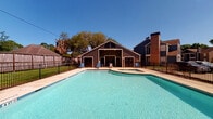 This single-story space is part of a residential community distinguished by its gray lap-siding exterior and straightforward, functional design. The unit's entry is accessed via a covered patio area, which provides a sheltered outdoor space outfitted with a glass-top table and lounge seating—a practical spot for relaxing or dining outdoors. The black entry door with a brass knob opens directly into the unit, while the covered overhang offers a degree of privacy and protection from the elements.

A standout feature of this property is its access to a generously sized community swimming pool and adjoining spa. The pool area is enclosed by a wrought-iron perimeter fence, ensuring security while maintaining open sightlines to the surrounding landscaped grounds and mature trees. The concrete pool deck is furnished with additional lounge chairs, and safety equipment is visibly mounted nearby. Under clear blue skies, the pool area receives abundant natural sunlight, enhancing its appeal as an outdoor amenity.

The broader community setting includes multi-story neighboring buildings, palm trees, and well-maintained green spaces visible beyond the pool fence, contributing to a warm, suburban atmosphere. Overall, the space offers a combination of private outdoor living and shared recreational amenities within a well-established community environment.