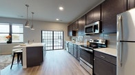 Located at 203 E Main St in Waunakee, Wisconsin, this contemporary single-floor unit offers a thoughtfully designed layout with modern finishes throughout. The kitchen serves as a central highlight, showcasing rich dark espresso cabinetry that extends to the ceiling, crisp white quartz countertops, and a full suite of stainless steel appliances including a range, built-in microwave, dishwasher, and refrigerator. A light gray subway tile backsplash adds a clean, polished touch, while a center island with pendant lighting provides additional prep space and casual seating. Light wood-tone luxury vinyl plank flooring flows throughout the main areas, complementing the neutral gray wall palette. Large windows and a glass-panel sliding door bring in generous natural light and connect the kitchen to the living area. The living space, visible in the floorplan, features a ceiling fan and ample room for comfortable furnishings. The bedroom is carpeted and cozy, with a horizontal window that allows in soft, diffused light. The bathroom is finished with dark vanity cabinetry, a white countertop, open shelving, and wood-look flooring, offering a clean and functional design. Overall, this unit presents a cohesive modern aesthetic with quality finishes in a prime Waunakee location.