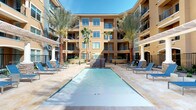 This expansive outdoor courtyard serves as a central amenity space defined by its resort-inspired design and sun-drenched atmosphere. At the heart of the space is a generously sized rectangular swimming pool with clear, blue-tinted water, surrounded by a travertine stone deck that provides a clean, upscale aesthetic underfoot. A dramatic stone-clad waterfall feature cascades into one end of the pool, adding both visual interest and ambient sound to the environment.

Blue chaise lounge chairs are arranged along the pool deck, offering ample space for sunbathing and relaxation. Shaded outdoor dining areas furnished with tables, chairs, and yellow market umbrellas provide comfortable spots for gathering away from direct sun. A white pergola structure near the waterfall adds an architectural focal point and additional shaded retreat.

The courtyard is enclosed on multiple sides by warm, earth-toned stucco buildings with balconies, large windows, and decorative stonework accents. Tall, mature palm trees are interspersed throughout, enhancing the tropical ambiance. Manicured desert landscaping and iron fencing complete the well-appointed outdoor setting, which benefits from abundant natural light under open skies.