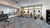 Located in Menomonee Falls, WI, this well-appointed fitness center offers a comprehensive workout environment across a single floor. The space is designed with functionality and comfort in mind, featuring a patterned, multi-tone carpet that adds visual interest while providing a durable surface underfoot. Recessed lighting and ceiling fans ensure the room remains bright and well-ventilated throughout.

The cardio zone is prominently positioned along a windowed wall, where a row of treadmills and elliptical machines face large windows fitted with adjustable blinds, allowing ample natural light to filter in. Each cardio unit is equipped with an integrated display screen for tracking workouts.

The strength training area is equally well-equipped, featuring a dual-cable functional trainer, a dumbbell rack with a full selection of free weights, and a weight bench. A large full-length mirror spans one wall, enhancing the sense of space and allowing users to monitor their form during exercise. A wall-mounted flat-screen television adds convenience for those looking to stay entertained.

Complementing the equipment is a dedicated rack stocked with medicine balls, stability balls, and kettlebells, supporting a wide range of functional and group fitness exercises. The overall ambiance is clean and modern, with yellow-green accent walls in the cardio section providing an energizing pop of color.