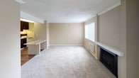 This single-floor space is organized into distinct functional areas connected by an open, practical layout. The living room is a generously sized, carpeted space finished in warm neutral tones with crown molding and a textured ceiling. A fireplace anchors one end of the room, while a large window with vertical blinds admits natural light and provides an outdoor view. A wall-mounted HVAC unit ensures year-round climate control, and a built-in ledge runs along one wall, adding architectural detail.

Adjacent to the living area, the kitchen features rich dark-stained cabinetry on both upper and lower levels, paired with light countertops and a classic white subway tile backsplash. A white drop-in sink, white electric coil-top range, and wood-look plank flooring complete the space, creating a practical and well-appointed cooking area.

A separate carpeted room, accessible from the main living space, offers a quiet and versatile area suitable for use as a bedroom or private retreat, with a pass-through view into the kitchen beyond.

The bathroom includes a full tub and shower combination with chrome fixtures, a toilet, and clean white finishes throughout, maintaining a simple and functional aesthetic consistent with the rest of the unit.