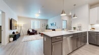 This single-story residence is designed with a clean, contemporary aesthetic that emphasizes open living and functional flow. The kitchen serves as the heart of the home, anchored by a large center island with a white quartz countertop and undermount sink. Gray cabinetry with brushed nickel hardware pairs with stainless steel appliances, including a full-size French door refrigerator and dishwasher. Pendant lighting in a matching brushed nickel finish adds a polished touch overhead. Light wood-look flooring extends seamlessly from the kitchen into the open living area, which is bright and welcoming, benefiting from natural light through windows fitted with horizontal blinds. The living space offers ample room for comfortable seating arrangements. The bedroom is a calm retreat featuring soft carpeting and a spacious walk-in closet equipped with a full-length hanging rod. The bathroom features a soaking tub with tile surround laid in a herringbone pattern, a dual-sink vanity with matching gray cabinetry and white countertops, and a large mirror. Throughout, the neutral color palette and consistent modern finishes create a cohesive and inviting atmosphere.
