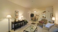 This single-floor residence offers an open-concept layout that connects the living and dining areas in a cohesive, flowing arrangement. The living area is anchored by a glass-top coffee table and neutral-toned upholstered seating, creating a relaxed and inviting atmosphere. Warm ambient lighting, provided by a combination of ceiling fixtures, a ceiling fan, and table lamps, casts a soft glow throughout the space, complementing the light-colored walls and neutral carpeted flooring.

A dark wood console table lines one wall, accessorized with framed artwork, a table lamp, and decorative greenery, adding visual depth and character. The adjoining dining area accommodates a dining table with wicker-style chairs, positioned beneath a ceiling fan with a built-in light kit. A patterned area rug beneath the dining set grounds the space and adds texture.

Built-in shelving is visible near what appears to be a kitchen pass-through area, offering practical display and storage options. Potted plants are thoughtfully placed throughout, bringing natural elements indoors and enhancing the overall warmth and livability of the space.