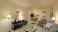 This single-floor residence offers an open-concept layout that connects the living and dining areas in a cohesive, flowing arrangement. The living area is anchored by a glass-top coffee table and neutral-toned upholstered seating, creating a relaxed and inviting atmosphere. Warm ambient lighting, provided by a combination of ceiling fixtures, a ceiling fan, and table lamps, casts a soft glow throughout the space, complementing the light-colored walls and neutral carpeted flooring.

A dark wood console table lines one wall, accessorized with framed artwork, a table lamp, and decorative greenery, adding visual depth and character. The adjoining dining area accommodates a dining table with wicker-style chairs, positioned beneath a ceiling fan with a built-in light kit. A patterned area rug beneath the dining set grounds the space and adds texture.

Built-in shelving is visible near what appears to be a kitchen pass-through area, offering practical display and storage options. Potted plants are thoughtfully placed throughout, bringing natural elements indoors and enhancing the overall warmth and livability of the space.
