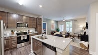 This thoughtfully designed 3-bedroom, 2-bathroom residence is laid out on a single floor, making efficient use of its space across a living room, kitchen, three bedrooms, two bathrooms, a hallway, and a closet. The open-concept kitchen and living area serves as the heart of the home, where rich dark-walnut cabinetry, granite-look countertops, a subway tile backsplash, and a full suite of stainless steel appliances — including a range, over-the-range microwave, refrigerator, and dishwasher — create a functional and stylish cooking environment. A central kitchen island with a double sink and bar seating bridges the kitchen and living space seamlessly. The living area is bright and welcoming, with large windows dressed in light curtains that invite natural light and offer outdoor views, complemented by wood-look flooring throughout. The primary bedroom features plush carpeting, natural light from a well-sized window, and ample room for furnishings. The additional bedrooms are similarly carpeted and comfortable. The primary bathroom offers a dual-sink vanity with granite-look countertops, wood-toned cabinetry, a large mirror, and wood-look tile flooring. The overall aesthetic is clean and contemporary, with neutral tones providing a versatile backdrop throughout.