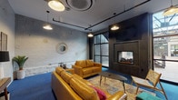 This single-floor amenity space combines industrial design with modern comfort across several distinct areas. The centerpiece is a stylish lounge that showcases exposed painted brick walls, bold blue carpeting, and a striking dark-tiled double-sided fireplace topped with a wall-mounted television. Large floor-to-ceiling windows flood the room with natural light and offer views of an adjacent outdoor terrace, while pendant lighting and black track fixtures overhead add a refined industrial character. Plush caramel leather sofas and armchairs, a patterned area rug, and eclectic accent pieces create a welcoming and vibrant gathering atmosphere. The fitness center is divided into two well-organized workout zones. The first zone houses treadmills, a dumbbell rack, and weight benches, while the second features cardio equipment including stationary bikes and an elliptical, a cable machine, and large wall mirrors that enhance the sense of space. Durable rubber flooring and bright linear lighting throughout make for a clean, functional training environment. The laundry facility is efficiently designed with rows of commercial-grade washers and dryers lining both walls, bright overhead lighting, and a neutral gray floor, ensuring a practical and tidy space for everyday convenience.