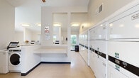 This shared laundry facility is a single-floor, open-plan space thoughtfully designed to accommodate a high volume of laundry needs efficiently. The room is outfitted with a substantial bank of top-loading washing machines along one side and a large row of stacked front-loading dryers along the opposite wall, providing ample capacity for simultaneous use. Structural columns divide the space into distinct zones without interrupting the overall open feel of the room.

A long, built-in granite-style folding counter runs centrally through the space, offering generous surface area for sorting and folding laundry. The machines feature modern card- and mobile payment-enabled controls, making transactions simple and cashless.

The space is finished with neutral, light-toned walls and durable tile flooring, contributing to a clean and functional aesthetic. Flush-mount ceiling light fixtures provide consistent overhead illumination, while multiple windows along the perimeter allow natural light to fill the room and offer views of surrounding greenery. The high ceilings near the entry enhance the sense of openness. A bulletin board near the entrance provides a space for notices and community information. The facility is monitored by a security surveillance system, ensuring a safe environment for all users.
