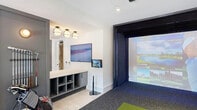 This thoughtfully designed golf simulator lounge offers a premier indoor golf experience within a contemporary, well-appointed space. At its centerpiece is a full enclosed simulator bay, complete with a large projection screen, impact-absorbing surround, and a bright green synthetic turf hitting mat — allowing users to virtually play renowned courses from around the world. Recessed and flush-mount lighting throughout ensures optimal visibility across all areas of the space.

The lounge area adjacent to the simulator bay features light wood-look flooring, an inviting arrangement of leather and upholstered seating, and large black-framed windows that allow generous natural light and outdoor views. A retro golf-themed arcade game adds a playful touch, complemented by curated wall art that reinforces the golf aesthetic.

A dedicated equipment zone includes a wall-mounted club rack and open-cubby storage cabinetry finished in a sleek gray, providing organized space for bags and gear. Multiple wall-mounted flat-screen displays offer additional viewing options and simulator controls. A separate corridor area features a built-in wet bar with a matte black faucet, white countertop, and bar seating, making it easy to entertain. The cohesive gray and white color palette, combined with warm accent lighting and stylish furnishings, gives the entire space a polished, upscale ambiance.
