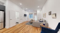 This well-appointed multi-floor space spans three levels, combining modern design with functional layouts throughout. Warm hardwood flooring flows consistently across each floor, providing a cohesive and inviting atmosphere. The main living area is generously proportioned, featuring recessed lighting, crisp white walls, and a wall-mounted television. A staircase connects the levels seamlessly, contributing to the open and airy feel of the interior.

The kitchen is a standout feature, outfitted with sleek charcoal cabinetry, a subway tile backsplash, stainless steel appliances including a full-size refrigerator, microwave, and range, along with a large island topped with a white stone countertop that doubles as a casual dining space with bar seating.

The bathroom is finished to a high standard, with floor-to-ceiling stone-effect tiles, a soaking tub with shower, a wide white vanity, and modern fixtures throughout. Additional rooms include a cozy bedroom with a natural light-admitting window, a dedicated office space furnished with a large desk and leather accent chairs, and a supplementary sitting area that offers flexible use.

Throughout the space, the design maintains a clean, contemporary aesthetic with neutral tones and thoughtful detailing, making it a well-rounded and highly functional environment across all three floors.