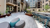 Located at 30 W Howell St in Boston's Dorchester neighborhood, this outdoor amenity courtyard is a thoughtfully designed communal space nestled within a modern multi-story residential building. The courtyard is framed on all sides by the building's contemporary façade, which features a warm combination of wood cladding, large windows, and private balconies that overlook the space below.

At the heart of the courtyard sits a resort-style swimming pool surrounded by a generous stone paver deck outfitted with lounge chairs, providing an inviting setting for relaxation. The pool area is complemented by glass panel enclosures and tall light poles that enhance both safety and ambiance.

The broader courtyard is finished with large-format gray stone pavers that create a clean, cohesive aesthetic throughout. Raised Corten steel planter beds are a defining architectural feature, filled with ornamental grasses, young trees, and lush greenery that add natural texture and warmth to the hardscaped environment.

Multiple seating zones cater to a range of social activities. A built-in granite bar with teal rope barstools serves as a casual gathering spot, while clusters of modern black chairs and tables provide dining options. Soft, rounded lounge poufs in gray and teal offer informal seating for relaxed conversation. A built-in gas grill station further enhances the space's outdoor entertaining capabilities, making this courtyard a well-appointed and functional outdoor retreat.