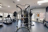 Located at 6565 McCallum Blvd in Dallas, TX, this fitness center offers a thoughtfully designed, single-floor workout space that accommodates a wide range of exercise needs. The layout is open and functional, with durable rubber flooring throughout that provides both safety and comfort underfoot.

The cardio zone features a row of equipment including treadmills, an elliptical, and a stationary bike, all positioned along a wall of large windows that flood the space with natural light and offer views to the outdoors. A wall-mounted television adds entertainment for longer cardio sessions, while a pair of leather lounge chairs provide a relaxed seating area nearby.

At the heart of the gym stands a robust Axis multi-functional cable and Smith machine system, fully loaded with weight plates and attachments for a comprehensive strength training experience. An adjustable weight bench and a full dumbbell rack round out the free weights section, catering to users of all fitness levels. Exercise and medicine balls are neatly stored on wall-mounted racks for easy access.

A striking navy blue accent wall, adorned with motivational fitness-themed typography in gold lettering, adds energy and personality to the space. Recessed lighting and skylights keep the entire area bright and inviting. A bulletin board near the entrance and a water station further enhance the user-friendly environment of this well-appointed community fitness center.