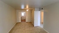 This single-floor residential space offers a straightforward and functional layout designed for comfortable everyday living. The main living area is finished with wall-to-wall neutral-toned carpeting, creating a warm and cohesive atmosphere throughout. A ceiling fan is centrally mounted, providing both practical air circulation and a subtle design element overhead. The space features smooth, neutral-painted walls with clean white baseboards that lend a simple, understated aesthetic. Built-in open shelving is integrated into the layout, offering convenient storage and display options without occupying additional floor space. Multiple doorways connect the primary living area to adjacent rooms, promoting a natural and accessible flow throughout the floor plan. One passageway leads to a room with hardwood-style flooring and a window that brings in natural light, brightening the interior. An overhead HVAC vent is visibly installed near the ceiling, indicating a central climate control system. The overall ambiance is clean, neutral, and move-in ready, with a practical design that prioritizes functionality and ease of use across the single level.