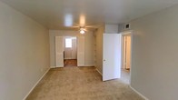 This single-floor residential space offers a straightforward and functional layout designed for comfortable everyday living. The main living area is finished with wall-to-wall neutral-toned carpeting, creating a warm and cohesive atmosphere throughout. A ceiling fan is centrally mounted, providing both practical air circulation and a subtle design element overhead. The space features smooth, neutral-painted walls with clean white baseboards that lend a simple, understated aesthetic. Built-in open shelving is integrated into the layout, offering convenient storage and display options without occupying additional floor space. Multiple doorways connect the primary living area to adjacent rooms, promoting a natural and accessible flow throughout the floor plan. One passageway leads to a room with hardwood-style flooring and a window that brings in natural light, brightening the interior. An overhead HVAC vent is visibly installed near the ceiling, indicating a central climate control system. The overall ambiance is clean, neutral, and move-in ready, with a practical design that prioritizes functionality and ease of use across the single level.
