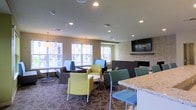 This single-floor communal space is designed as a stylish and functional lounge area, offering a welcoming environment for gathering and relaxation. The room is characterized by its modern, open layout with recessed lighting throughout the ceiling, providing consistent and even illumination across the space.

Seating is arranged in a variety of flexible groupings, featuring an eclectic mix of contemporary chairs in yellow-green, teal, and patterned upholstery, paired with round white accent tables that encourage casual conversation. Along the window-facing wall, fixed booth-style seating with individual tables offers a more private setting bathed in natural light from a row of large windows overlooking an outdoor area.

A standout feature of the room is the wall-mounted flat-screen television positioned above a stone-accented fireplace surround, creating a natural focal point that adds warmth and character. Adjacent to the seating area, a granite-topped counter surface suggests a refreshment or service station nearby. The patterned area rug grounds the seating arrangement and adds visual texture, while pendant lighting near the window wall contributes a refined finishing touch to the overall aesthetic.