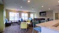 This single-floor communal space is designed as a stylish and functional lounge area, offering a welcoming environment for gathering and relaxation. The room is characterized by its modern, open layout with recessed lighting throughout the ceiling, providing consistent and even illumination across the space.

Seating is arranged in a variety of flexible groupings, featuring an eclectic mix of contemporary chairs in yellow-green, teal, and patterned upholstery, paired with round white accent tables that encourage casual conversation. Along the window-facing wall, fixed booth-style seating with individual tables offers a more private setting bathed in natural light from a row of large windows overlooking an outdoor area.

A standout feature of the room is the wall-mounted flat-screen television positioned above a stone-accented fireplace surround, creating a natural focal point that adds warmth and character. Adjacent to the seating area, a granite-topped counter surface suggests a refreshment or service station nearby. The patterned area rug grounds the seating arrangement and adds visual texture, while pendant lighting near the window wall contributes a refined finishing touch to the overall aesthetic.