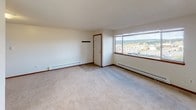 Located at 19630 Ashcrest Loop in Poulsbo, Washington, this single-floor unit offers a straightforward and functional layout designed for comfortable everyday living. The living area is generously sized, finished with plush wall-to-wall carpeting and neutral-toned walls that create a clean, versatile backdrop. Baseboard heating runs along the perimeter, and a large window frames views of the surrounding neighborhood and tree-lined landscape, filling the space with natural light. The kitchen is efficiently arranged with warm honey-toned wood cabinetry, white appliances including a refrigerator, range, and dishwasher, and tile flooring that transitions seamlessly from the countertops to the work area. Ample upper and lower cabinet storage provides practical utility. A partial wall separates the kitchen from the main living space, maintaining an open feel while defining distinct functional zones. The bathroom is well-appointed with a full soaking tub and shower combination, a wood-framed vanity with a single sink, and tile flooring throughout. The space maintains a consistent design language with warm wood accents and light neutral tones. Overall, this unit presents a practical and well-maintained living environment in the Poulsbo community.