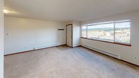 Located at 19630 Ashcrest Loop in Poulsbo, Washington, this single-floor unit offers a straightforward and functional layout designed for comfortable everyday living. The living area is generously sized, finished with plush wall-to-wall carpeting and neutral-toned walls that create a clean, versatile backdrop. Baseboard heating runs along the perimeter, and a large window frames views of the surrounding neighborhood and tree-lined landscape, filling the space with natural light. The kitchen is efficiently arranged with warm honey-toned wood cabinetry, white appliances including a refrigerator, range, and dishwasher, and tile flooring that transitions seamlessly from the countertops to the work area. Ample upper and lower cabinet storage provides practical utility. A partial wall separates the kitchen from the main living space, maintaining an open feel while defining distinct functional zones. The bathroom is well-appointed with a full soaking tub and shower combination, a wood-framed vanity with a single sink, and tile flooring throughout. The space maintains a consistent design language with warm wood accents and light neutral tones. Overall, this unit presents a practical and well-maintained living environment in the Poulsbo community.