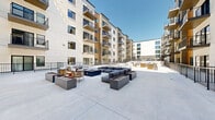 Located at 3078 Decker Lake Dr in West Valley City, Utah, this outdoor amenity courtyard is a well-appointed communal space set at the ground level of a contemporary multi-story residential complex. The courtyard features a clean, modern design with a spacious concrete patio that accommodates multiple distinct seating and gathering areas. A central lounge zone anchors the space with a large sectional sofa arrangement surrounding a rectangular fire pit table, providing a comfortable setting for socializing. A second seating area nearby features woven rattan lounge chairs with plush white cushions positioned around a cylindrical fire pit, offering a more intimate atmosphere. Raised rectangular planters in a neutral tone are integrated throughout, adding architectural structure to the outdoor layout. The courtyard is enclosed by sleek black metal railings and bordered on multiple sides by the multi-story building facade, which features a modern exterior with a mix of white panel cladding and warm wood-tone accents. Large windows and private balconies line the upper floors, overlooking the courtyard below. String light fixtures are suspended overhead, enhancing the ambiance during evening hours. The open-air design allows for abundant natural light throughout the day, making it a versatile space suited for both relaxed and social use.